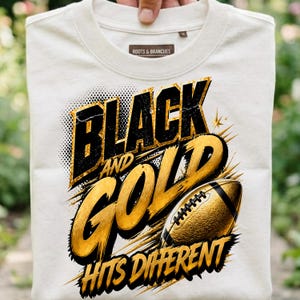 May include: A cream-colored t-shirt featuring a graphic design with the text "BLACK AND GOLD HITS DIFFERENT" in bold black and gold lettering. The design includes a textured gold football illustration with black accents and dynamic motion lines.