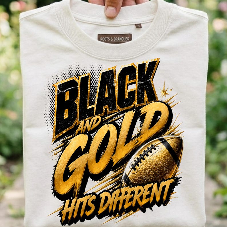 May include: A cream-colored t-shirt featuring a graphic design with the text "BLACK AND GOLD HITS DIFFERENT" in bold black and gold lettering. The design includes a textured gold football illustration with black accents and dynamic motion lines.