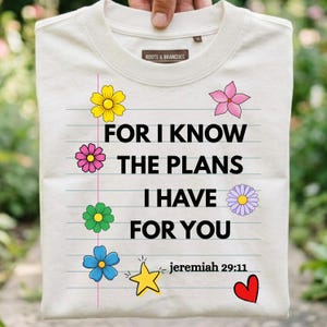 Peut inclure: Un t-shirt couleur crème avec un motif de papier ligné portant le texte "FOR I KNOW THE PLANS I HAVE FOR YOU" et "jeremiah 29:11". Le design comprend des illustrations colorées de fleurs, une étoile jaune et un cœur rouge.