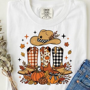 Peut inclure: Un t-shirt blanc avec une impression graphique représentant un chapeau de cowboy beige, trois bottes de cowboy à motifs carreaux orange, citrouilles et carreaux noirs, entourées de feuilles d'automne, de glands et d'étoiles noires.