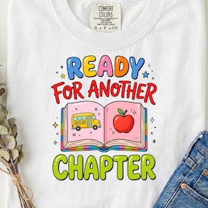 May include: A white t-shirt featuring a colourful graphic print. The text reads READY FOR ANOTHER CHAPTER in blue, red, and green. The design includes an open book with a yellow school bus on the left page and a red apple on the right page.