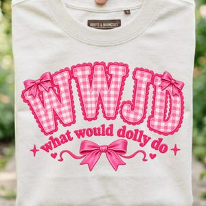 Peut inclure: Un t-shirt couleur crème arborant le texte WWJD dans un motif vichy rose et blanc avec des bords festonnés et des nœuds roses. En dessous, le texte what would dolly do est imprimé en rose, agrémenté d'un grand nœud rose, de cœurs et d'étoiles.