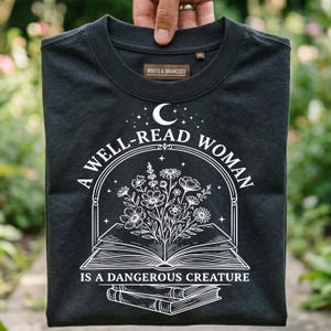 Peut inclure: Un t-shirt noir à col rond avec une impression graphique blanche représentant un livre ouvert d'où sortent des fleurs, surmonté d'un croissant de lune et d'étoiles, avec le texte A WELL-READ WOMAN IS A DANGEROUS CREATURE imprimé autour.
