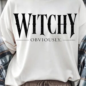 May include: A cream-colored crewneck sweatshirt featuring the black text WITCHY OBVIOUSLY printed across the chest in a bold, serif font. The word WITCHY is in a large, distressed typeface, while OBVIOUSLY is in a smaller, clean font with lines.
