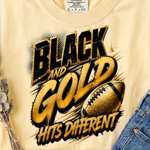 May include: A pale yellow t-shirt featuring a graphic design with the text "BLACK AND GOLD HITS DIFFERENT" in bold black and gold lettering. The design includes a textured gold football illustration with black laces and dynamic starburst accents.
