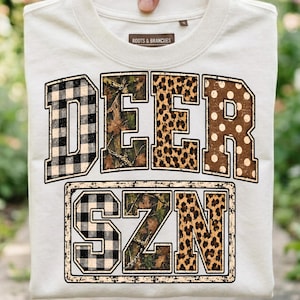 May include: A cream-coloured t-shirt featuring the text DEER SZN in large block letters. The letters are filled with various patterns including black and white buffalo plaid, camouflage, leopard print, and brown with white polka dots.