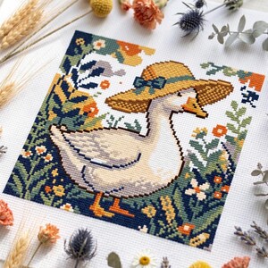 May include: A cross-stitch embroidery pattern on white fabric featuring a white goose wearing a wide-brimmed yellow straw hat with a teal ribbon. The goose is surrounded by a dense garden of dark blue foliage, orange flowers, and small white blossoms.