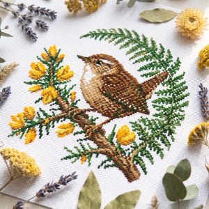 Puede incluir: Un bordado de punto de cruz sobre tela blanca que muestra un pájaro marrón posado en una rama con flores amarillas y follaje verde tipo helecho. El diseño está rodeado de lavanda seca, flores amarillas y hojas de eucalipto.