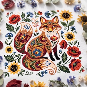 May include: A cross-stitch embroidery on white fabric featuring a central fox decorated with intricate floral patterns in shades of orange, red, blue, and yellow, surrounded by a border of sunflowers, red poppies, and blue cornflowers.