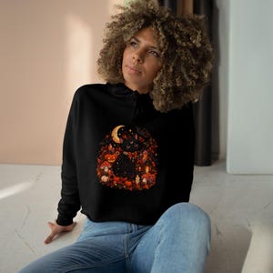 Peut inclure: Un sweat à capuche court noir avec une impression graphique centrale représentant un chat noir entouré d'éléments d'automne, notamment des citrouilles orange, des champignons rouges, des feuilles mortes, un croissant de lune et des lanternes.