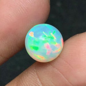 Könnte beinhalten: Ein runder, schillernder Opal-Edelstein. Der Opal zeigt ein lebendiges Farbspiel mit Blitzen in Blau, Grün, Rosa und Orange. Der Edelstein wird zwischen zwei Fingern gehalten, was seine kugelförmige Form und seine leuchtende Qualität hervorhebt.
