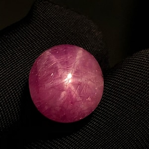 Peut inclure: Une pierre précieuse rubis étoilé de forme ronde et taillée en cabochon, d'une teinte magenta-rose profond. La pierre présente un effet d'étoile à six branches distinct sur sa surface polie et translucide, avec des inclusions internes.