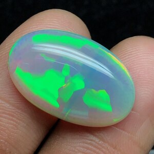 Könnte beinhalten: Ein ovaler Opal-Cabochon-Edelstein mit einer durchscheinenden weißen Basis und leuchtend grünen Farbblitzen auf der Oberfläche. Der Stein hat eine glatte, polierte und gewölbte Oberseite, die das Licht mit einem hellen Glanz reflektiert.