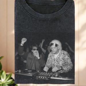 May include: A charcoal gray crew neck t-shirt featuring a black and white graphic print of two individuals at a DJ booth. One person wears sunglasses and has a hand raised, while the other wears a metallic headband and a patterned long-sleeve shirt.
