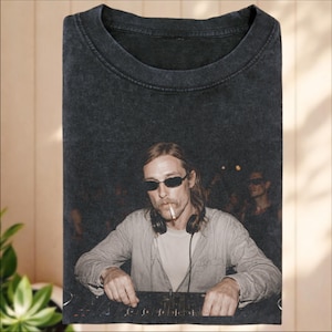 Pode incluir: Uma t-shirt cinzento-carvão com efeito de lavagem ácida, apresentando uma impressão gráfica de uma pessoa de cabelo comprido, óculos de sol escuros e auscultadores, a fumar um cigarro enquanto opera uma mesa de mistura de DJ.