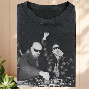 Może przedstawiać: Czarny t-shirt z efektem sprania, ozdobiony czarno-białym nadrukiem graficznym przedstawiającym dwie osoby w okularach przeciwsłonecznych za mikserem DJ-skim. Jedna osoba ma ogoloną głowę i bródkę, a druga ma otwarte usta i nosi koszulę w kratę.