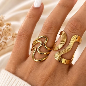May include: Two gold-colored rings with unique wave and curved designs. The rings are worn on a hand with white nail polish. The rings have a shiny, metallic finish and are likely made of metal.