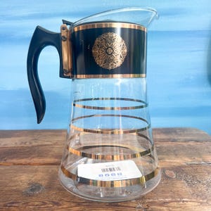 May include: A clear glass pitcher featuring a black band at the top with a gold circular filigree design. The body of the pitcher is decorated with horizontal gold stripes. It includes a black handle and a metal hinge mechanism at the top.