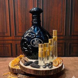 May include: A large black glass perfume bottle labeled NYLA SUEDE with three smaller clear glass spray vials labeled Handcrafted by Smoke & Oud 1ML, 2ML, and 5ML. The items are arranged on a wooden slice with dried orange peels and dark seeds.