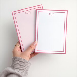 May include: Two personalized notepads with pink borders and the initials "K.C." printed in the upper center. One notepad is held in a hand, displaying a light pink background, while the other has a white background.