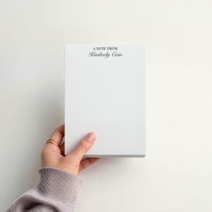 May include: A white notepad held by a hand, with the text "A Note From Kimberly Cain" printed in black at the top. The notepad is rectangular and appears to be made of thick paper. The background is a plain white surface.