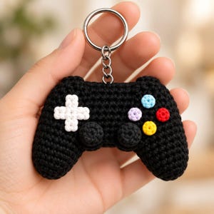 Könnte beinhalten: Ein schwarzer gehäkelter Schlüsselanhänger in Form eines Videospiel-Controllers. Der Controller verfügt über ein weißes kreuzförmiges Steuerkreuz auf der linken Seite und vier kleine, runde Knöpfe in Hellblau, Lavendel, Rot und Gelb rechts.