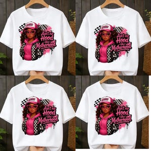 Può includere: T-shirt bianche con una grafica raffigurante una donna con una felpa con cappuccio rosa, una giacca a scacchi e un berretto. Il testo "Ain't no Hood like Motherhood" è visualizzato con accenti rosa e un design a bandiera a scacchi.