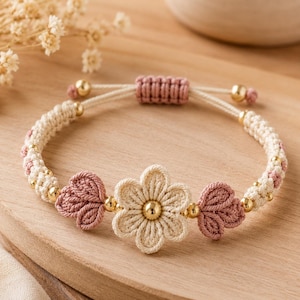 May include: A macrame bracelet featuring a central cream-colored flower with a gold bead center, flanked by two dusty pink leaf-shaped knots. The band consists of cream and pink woven cord, accented with small gold beads and a sliding knot closure.