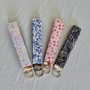May include: Four fabric key fobs in a row, each with a different pattern. The patterns include floral designs in pink, blue, and a black and white paisley. Each fob has a metal ring and a matching metal end.