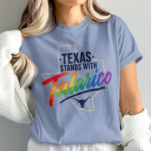 Könnte beinhalten: Ein hellblaues Kurzarm-T-Shirt mit einer Umrissgrafik des Bundesstaates Texas. Der Text "TEXAS STANDS WITH" ist in Dunkelblau gedruckt, darunter steht das Wort "Talarico" in Regenbogenfarben, ergänzt durch ein dunkelblaues Longhorn-Symbol.