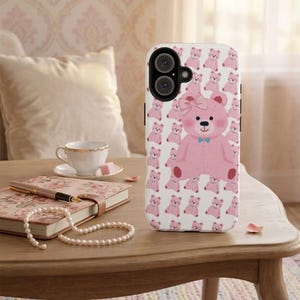 Peut inclure: Coque de téléphone blanche avec un motif répété d'ours en peluche roses. Un grand ours en peluche rose avec un nœud et un nœud papillon bleu est centré sur la coque. La coque est sur une table en bois avec un journal, un stylo et un collier de perles.