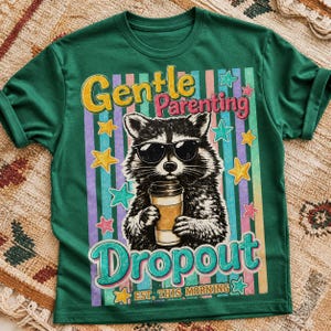 Puede incluir: Camiseta verde con un diseño gráfico de un mapache con gafas de sol sosteniendo una taza de café. La camiseta tiene las palabras "Gentle Parenting Dropout" y "Est. This Morning" en una fuente retro.