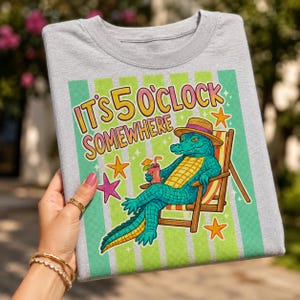May include: A grey t-shirt with the words "IT'S 5 O'CLOCK SOMEWHERE" in yellow and pink. The shirt features a cartoon alligator in a beach chair, wearing a hat and holding a drink. The design includes stars and green and white stripes.