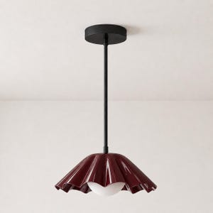 Op de afbeelding: Een hanglamp met een dieprode, glanzende, geplooide kap met een geschulpte rand. De kap hangt aan een dunne, matzwarte metalen stang die is verbonden met een bijpassende ronde zwarte plafondkap, voorzien van een witte bolvormige lamp.