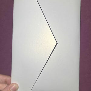 Peut inclure: Une enveloppe rectangulaire blanche avec un rabat triangulaire pointu. Le papier présente une finition nacrée et légèrement scintillante qui reflète la lumière, créant un éclat doux le long des bords du rabat et du corps de l'enveloppe.