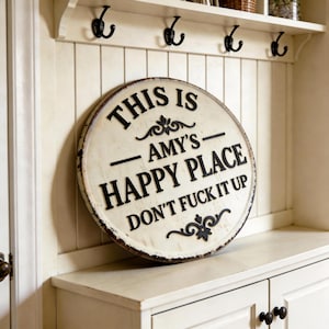 Könnte beinhalten: Ein rundes, cremefarbenes Wandschild mit abgenutzter Optik und dunklen Rändern. Das Schild trägt erhabene schwarze Schriftzüge mit dem Text: THIS IS AMY'S HAPPY PLACE DON'T FUCK IT UP, ergänzt durch zwei dekorative schwarze Schnörkel-Verzierungen.