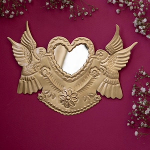 Op de afbeelding: Een goudkleurige metalen wandspiegel in de vorm van een hart, geflankeerd door twee symmetrische vogels met gespreide vleugels. Het frame is voorzien van reliëf bloemmotieven en geschulpte randen rondom een hartvormig spiegelend glasoppervlak.