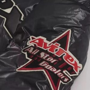 Pode incluir: Close-up de um casaco preto com acabamento brilhante. O casaco apresenta um patch de estrela bordado em vermelho e branco com o texto "Avirex All Star Dealers". O casaco tem patches brancos e pretos.