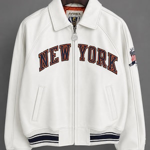 May include: White leather jacket with the text "NEW YORK" in navy and orange lettering across the chest. The jacket has a zip-up front, a collar, and navy and white striped cuffs and hem. An American flag patch is on the sleeve.