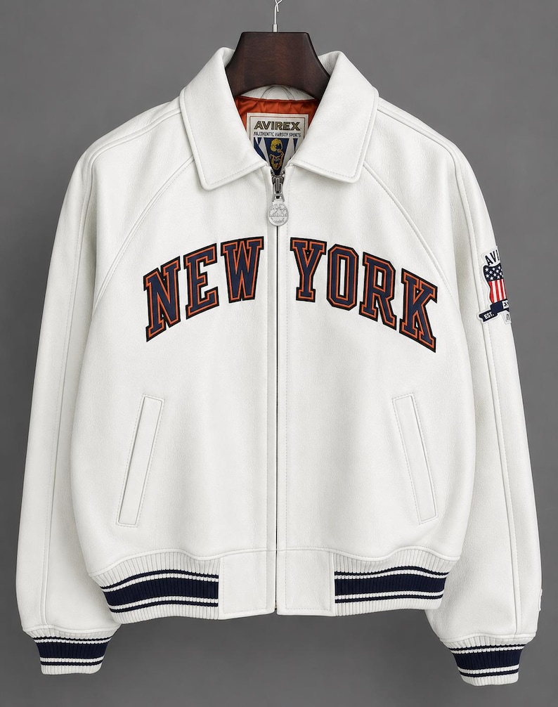 May include: White leather jacket with the text "NEW YORK" in navy and orange lettering across the chest. The jacket has a zip-up front, a collar, and navy and white striped cuffs and hem. An American flag patch is on the sleeve.