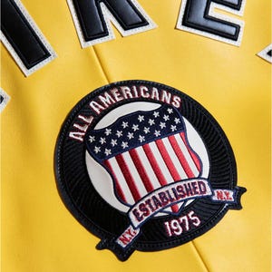 Pu&ograve; includere: Primo piano di una giacca gialla con una toppa circolare. La toppa presenta un disegno a scudo con strisce rosse e bianche, stelle blu e le parole "ALL AMERICANS ESTABLISHED 1975 N.Y." in lettere bianche. In alto sono visibili lettere nere e bianche.