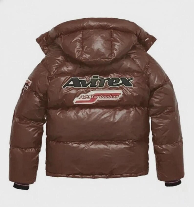 Pode incluir: Casaco puffer castanho com capuz. O casaco apresenta o nome da marca "Avirex" em letras brancas e pretas nas costas, com "All Star Dealers" por baixo. O casaco tem um acabamento brilhante e um design acolchoado.