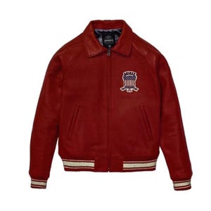 May include: A burgundy leather bomber jacket with a zip-up front, collar, and ribbed cuffs and hem. The jacket features a patch on the chest with an emblem and the brand name. The cuffs and hem have white stripes.