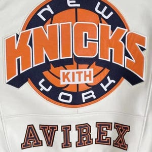 May include: Cream-colored leather jacket featuring the New York Knicks logo in orange and navy blue. The jacket includes the text "KITH" and "AVIREX" in a contrasting color. The jacket has a striped navy blue and white trim.
