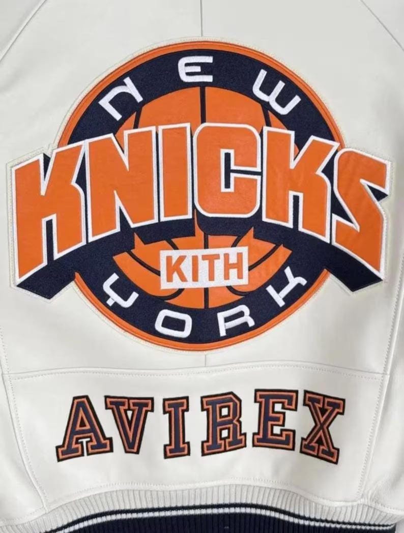 May include: Cream-colored leather jacket featuring the New York Knicks logo in orange and navy blue. The jacket includes the text "KITH" and "AVIREX" in a contrasting color. The jacket has a striped navy blue and white trim.