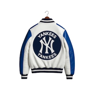 Op de afbeelding: Witte en blauwe Yankees honkbaljas met een marineblauwe ronde patch op de rug. De patch toont het logo van het team en het woord "Yankees" in witte letters. De jas heeft blauwe mouwen en een gestreepte kraag en manchetten.