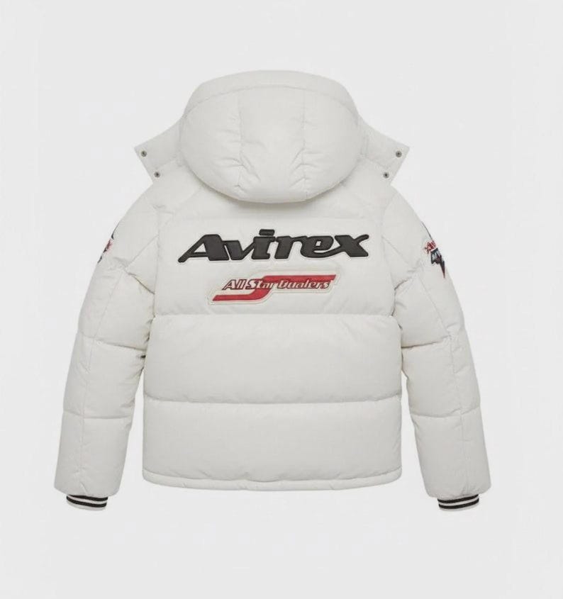 Pode incluir: Casaco acolchoado branco com capuz. O casaco apresenta o nome da marca "Avirex" em letras pretas nas costas, com "All Star Leaders" por baixo. O casaco tem punhos listrados em preto e branco e patches nas mangas.