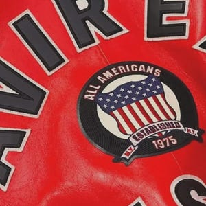 Pu&ograve; includere: Primo piano di una giacca di pelle rossa con scritte bianche e nere. Una toppa circolare presenta la bandiera americana e le parole "ALL AMERICANS ESTABLISHED 1975 N.Y."