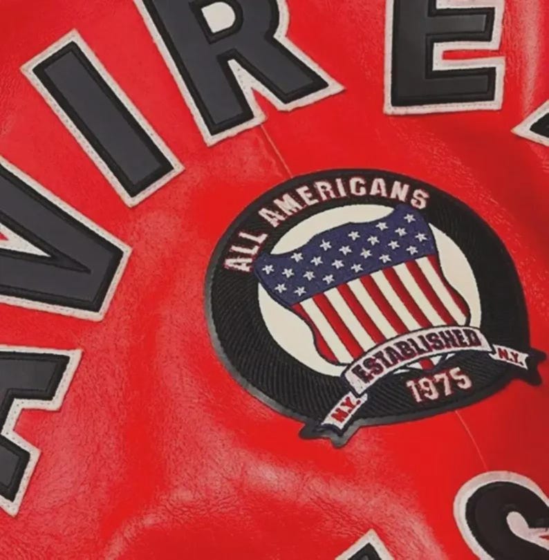Pu&ograve; includere: Primo piano di una giacca di pelle rossa con scritte bianche e nere. Una toppa circolare presenta la bandiera americana e le parole "ALL AMERICANS ESTABLISHED 1975 N.Y."