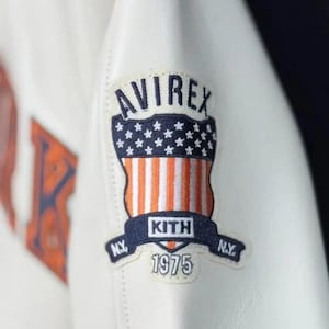 May include: Close-up of a white leather jacket featuring a patch with the "AVIREX" logo above a design of stars and stripes. Below the design is a banner with "KITH" and "1975" printed on it. The patch also has "N.Y." on each side.